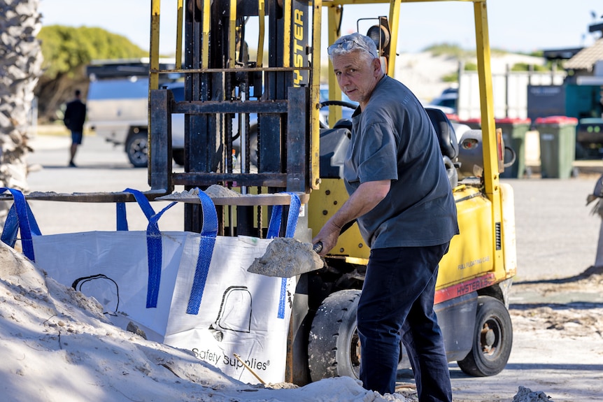 Glen Trebilcock lifts a shovel full of sand, in front of a fork lift holding an open sand bag 