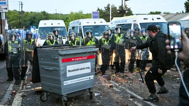 A line of police officers wearing riot gear are standing, facing a protester wearing all black and holding what appears to be a selfie stick. There is a metal commercial bin in front of the officers