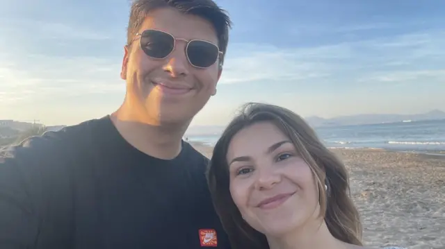 Young man in blue t-shirt with a red Nike logo, wearing sunglasses stands next to young woman wearing a floral top, with a beach in the background