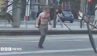 A still from a video showing a bare-chested man wearing dark trousers standing in the middle of a street. He is carrying a weapon in his hand. The still is blurry and the man's face has been delibrately blurred.