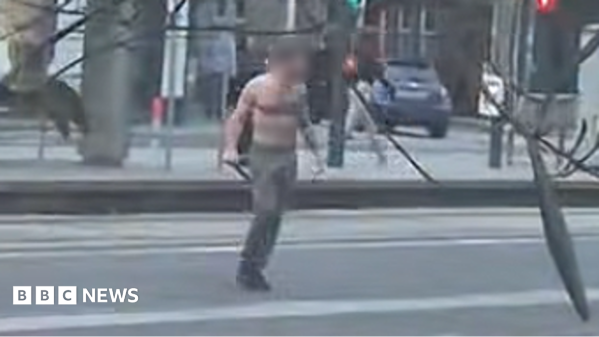 A still from a video showing a bare-chested man wearing dark trousers standing in the middle of a street. He is carrying a weapon in his hand. The still is blurry and the man's face has been delibrately blurred.