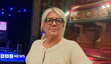 A woman with blonde hair and glasses stands in an empty theatre.