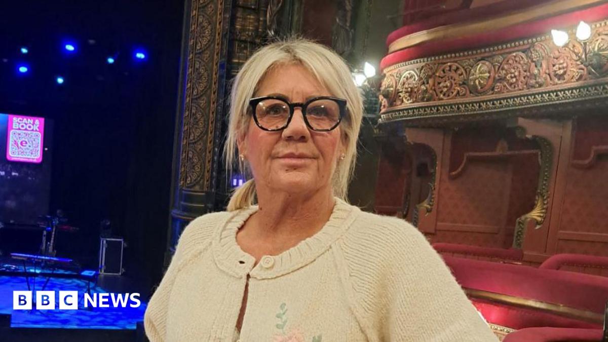 A woman with blonde hair and glasses stands in an empty theatre.