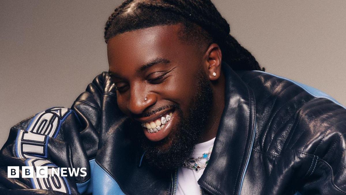 Mark Orabiyi looking down smiling. He is wearing a black and blue leather jacket over a white T-shirt