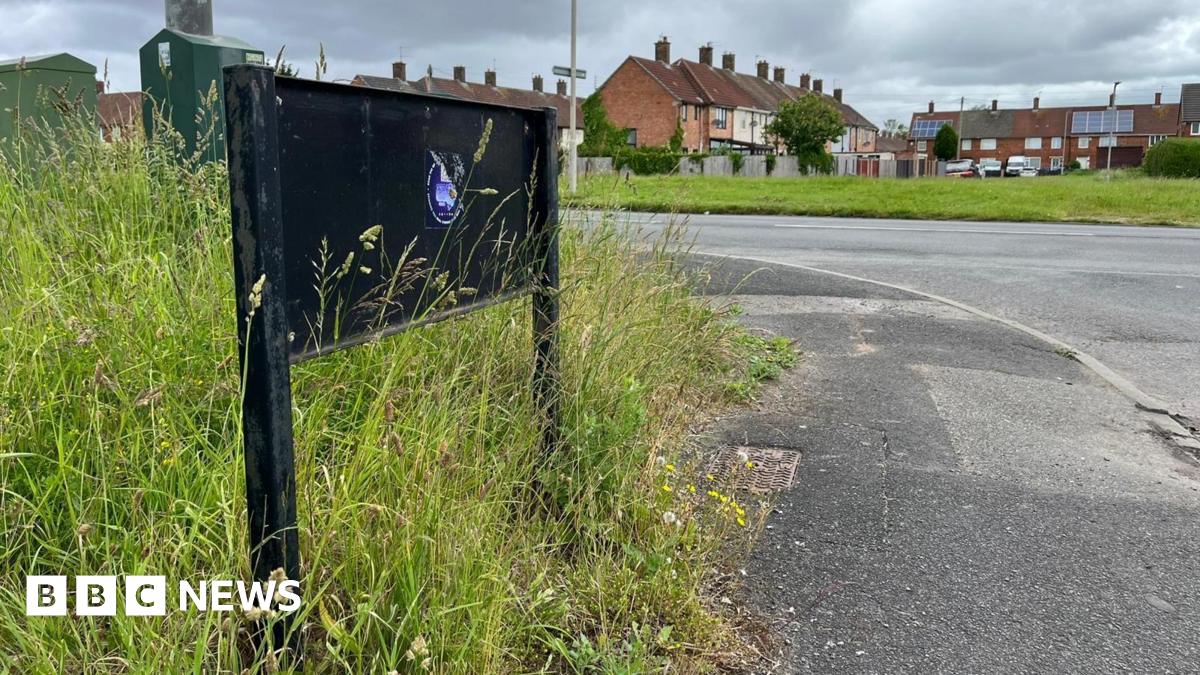 A black metal frame where the Dungeon Lane road sign would have been is surrounded by long grass. A main road runs alongside the lane with an terraced houses in the background.