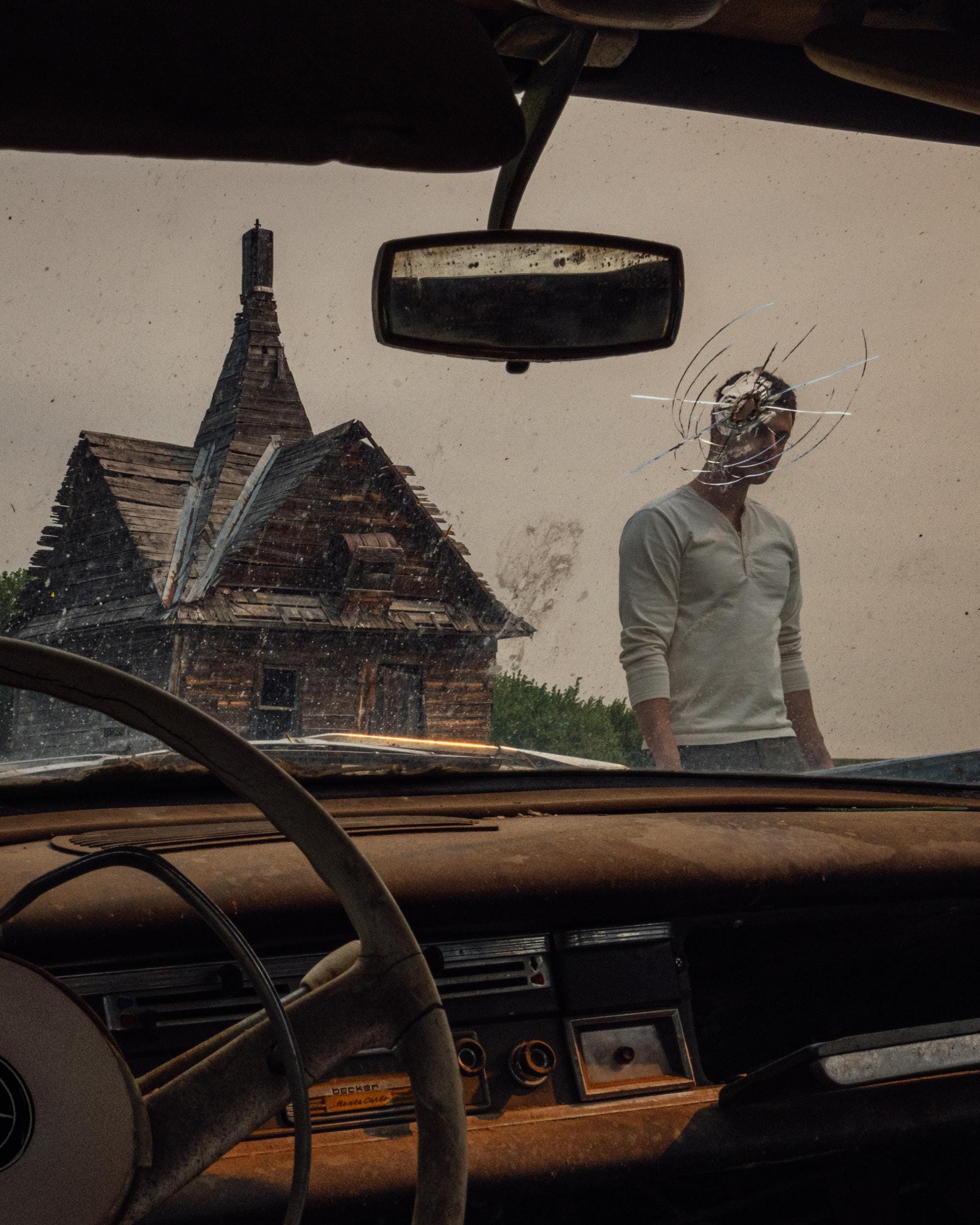 a photograph by Brendon Burton from the driver's seat inside a car. a man stands in front of the car, his head lined up visually with a bullet hole in the windshield. a derelict house stands in the background