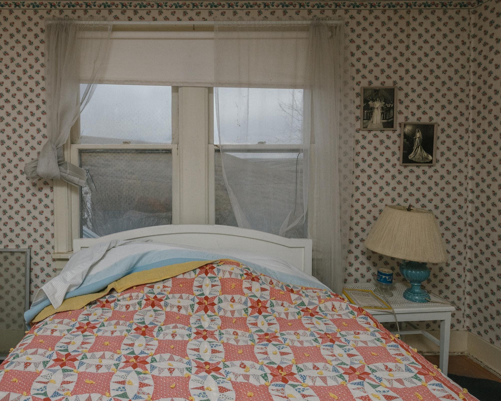 a photograph by Brendon Burton of a bed, nightstand, lamp, and two window panes in a bedroom