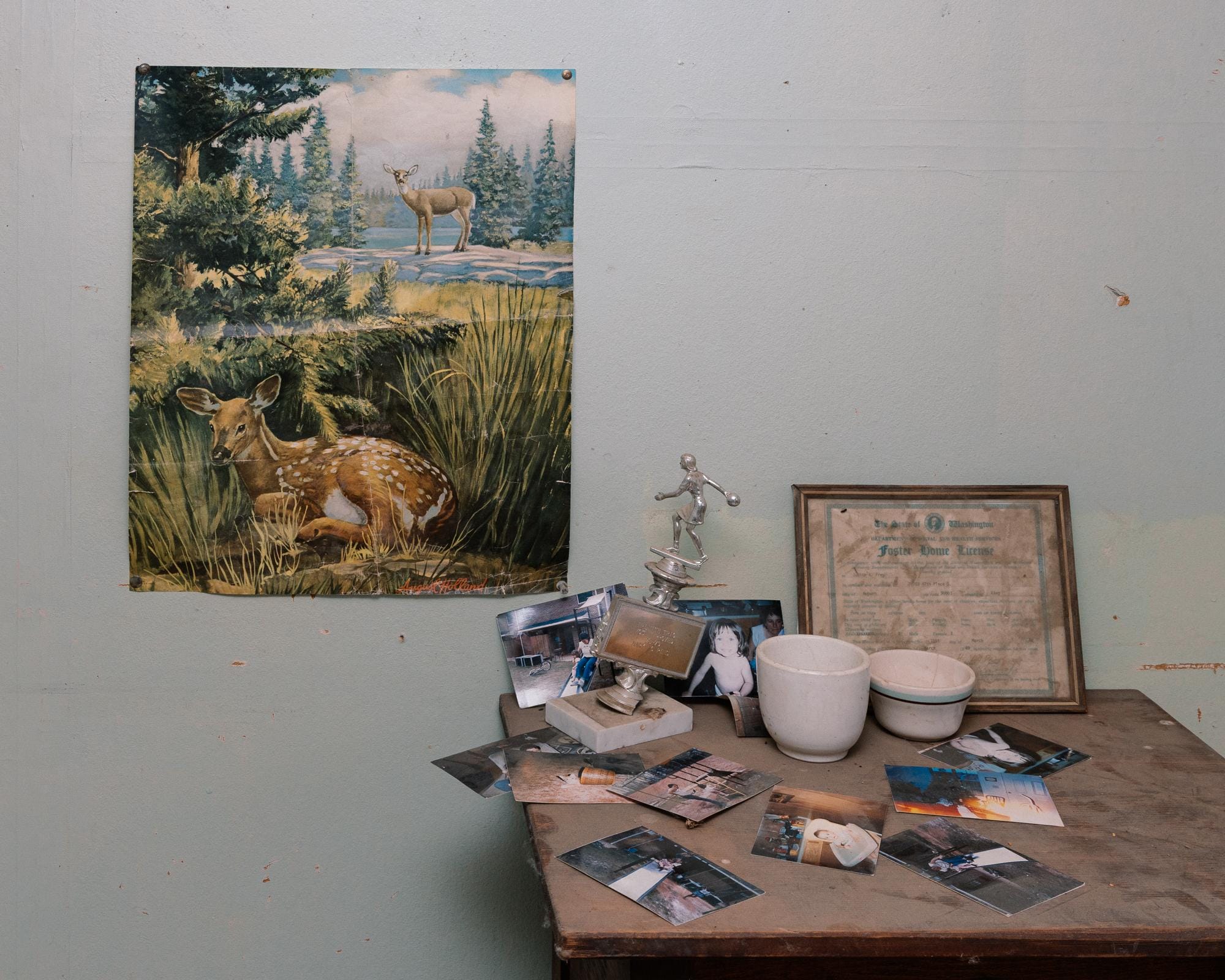 a photograph by Brendon Burton of a poster of deer pinned to the wall next to a table of a framed Foster Home License, photos, an old trophy, and mugs.