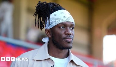 KSI wears a tan shirt over a white t-shirt. He has black dreadlocks and wears a white bandana around his forehead.