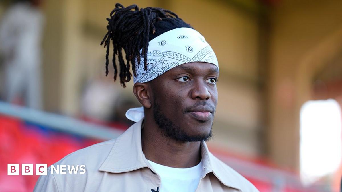 KSI wears a tan shirt over a white t-shirt. He has black dreadlocks and wears a white bandana around his forehead.