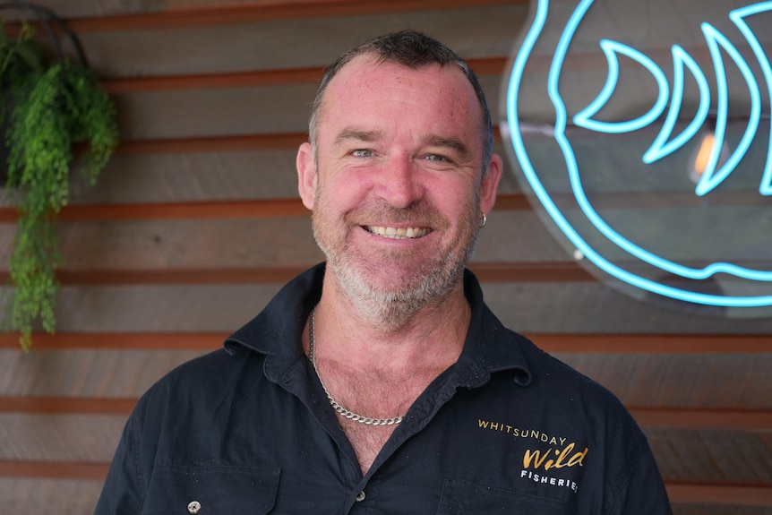 A man with short, dark hair wears a branded shirt and smiles as he stands in front of a neon sign in a restaurant.