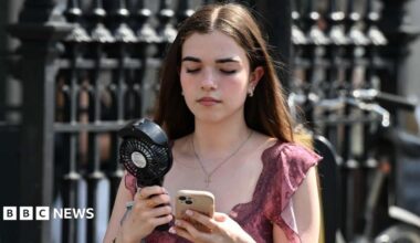 A young girl in a pink top with brown hair walks along a street carrying a portable black fan, whilst looking down at her phone in a clear case.