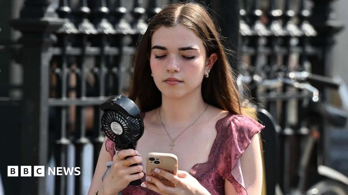 A young girl in a pink top with brown hair walks along a street carrying a portable black fan, whilst looking down at her phone in a clear case.