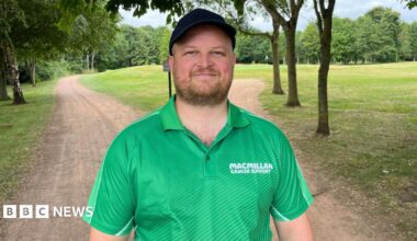 A man stands under trees on a dirt track on a golf course. He is wearing a bright green Macmillan Cancer Support polo shirt and a black cap. He has a short brown beard and he is smiling.