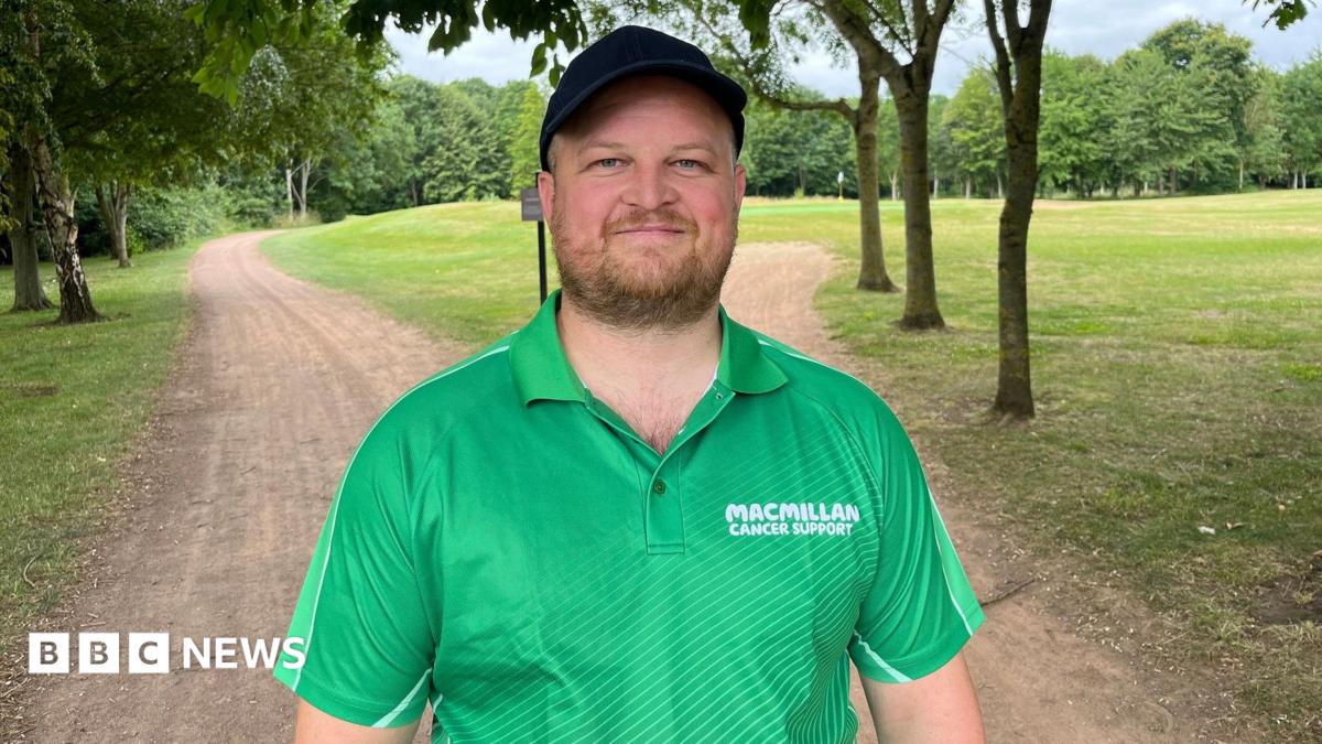 A man stands under trees on a dirt track on a golf course. He is wearing a bright green Macmillan Cancer Support polo shirt and a black cap. He has a short brown beard and he is smiling.