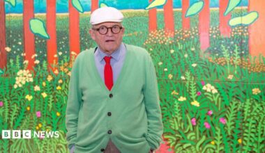 Hockney in a green cardigan, blue shirt, and orange tie in front of one of his paintings