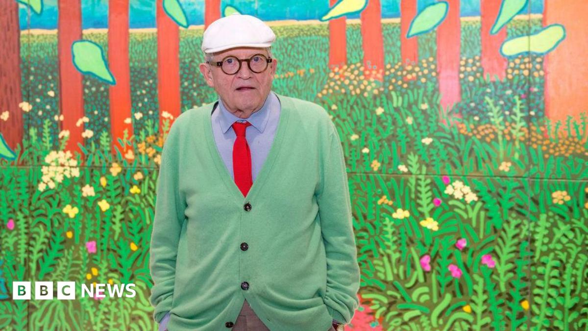Hockney in a green cardigan, blue shirt, and orange tie in front of one of his paintings