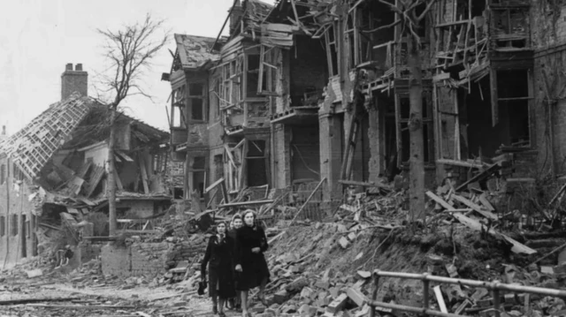 A picture of building destroyed during World War Two. Three people can be seen walking in front of the rubble.