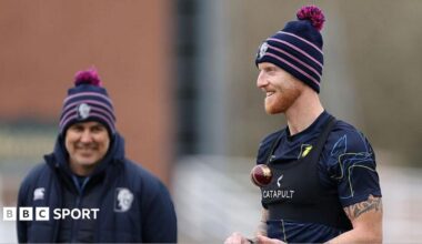 Ben Stokes in T-shirt and woolly hat, throws a red cricket ball up as he prepares to bowl in training as coach Ryan Campbell, in navy jacket and woolly hat watches on from a slight distance