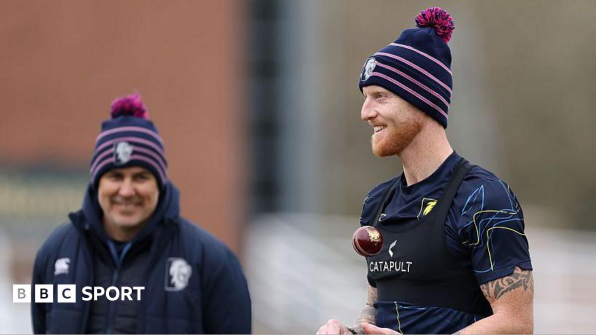 Ben Stokes in T-shirt and woolly hat, throws a red cricket ball up as he prepares to bowl in training as coach Ryan Campbell, in navy jacket and woolly hat watches on from a slight distance