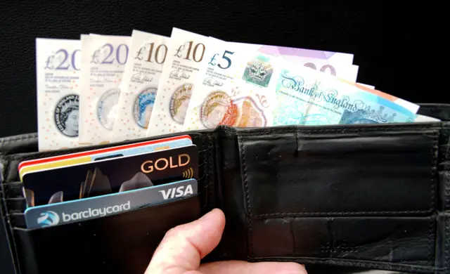 A five pound note, two ten pound notes and two twenty pound notes poke out of a black leather waller that is also holding several credit cards in the front