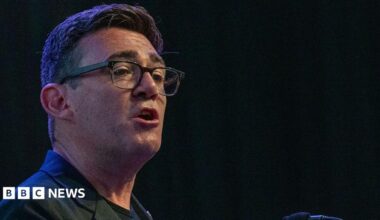 Andy Burnham wearing a black jacket with a black t-shirt talking to an audience with a black background behind him