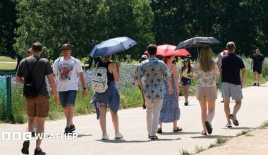 people walking on a pathway through a park.  Some are holding umbrellas to shade them from the hot weather