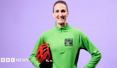 Jill Scott is standing in front of a purple background. She is smiling widely as she holds a red helmet under her arm. She is wearing a green quarter-zip top with Jill Scott's Coming Home Challenge written on it. Her hair is slicked back in a pony tail.
