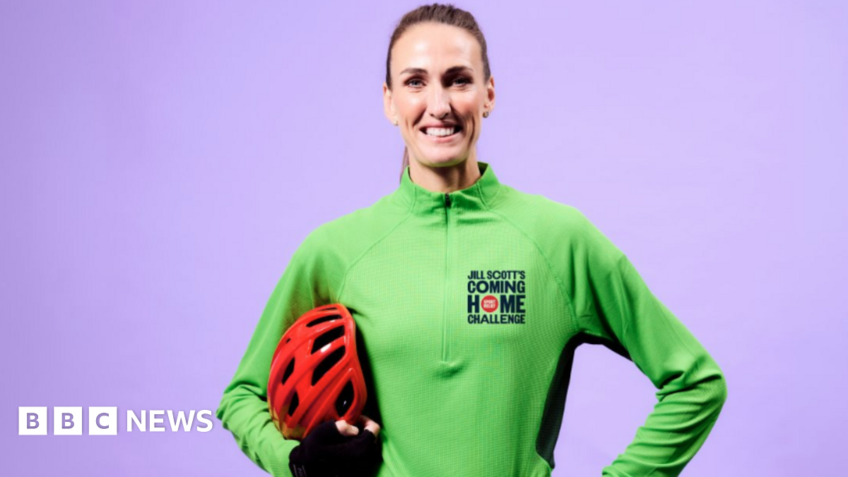 Jill Scott is standing in front of a purple background. She is smiling widely as she holds a red helmet under her arm. She is wearing a green quarter-zip top with Jill Scott's Coming Home Challenge written on it. Her hair is slicked back in a pony tail.