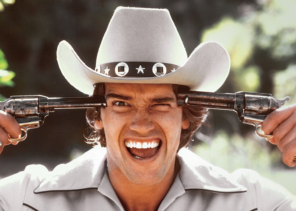 A color close-up of Schwarzenegger in a cowboy hat, laughing broadly while holding two revolvers level with his temples, their barrels pointed outward on either side of his head.