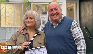 Ruth Jones stands with Steve Speirs with his arm around her. She is smiling holding a clapperboard, wearing a brown jacket has has a short grey bob. Steve Speirs is smiling, with a checked shirt with a blue jumper vest over it. A shop background is behind them, with a box of peppers in sight.