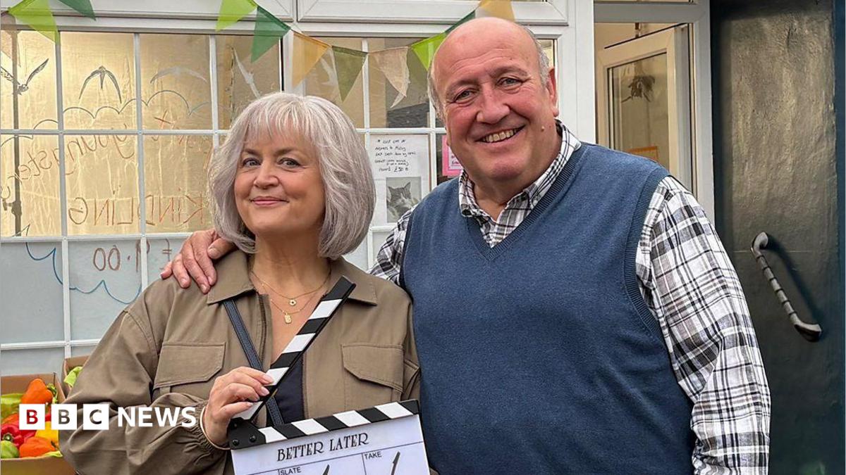 Ruth Jones stands with Steve Speirs with his arm around her. She is smiling holding a clapperboard, wearing a brown jacket has has a short grey bob. Steve Speirs is smiling, with a checked shirt with a blue jumper vest over it. A shop background is behind them, with a box of peppers in sight.