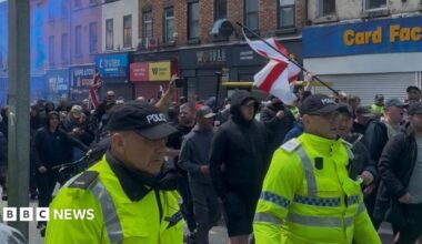 A large group of protesters, flanked by police officers in reflective yellow jackets, march along a street lined with shops, all of which have their shutters pulled down. Some protesters are carrying English and British flags, and some blue smoke from a flare is visible hanging in the air to the left.