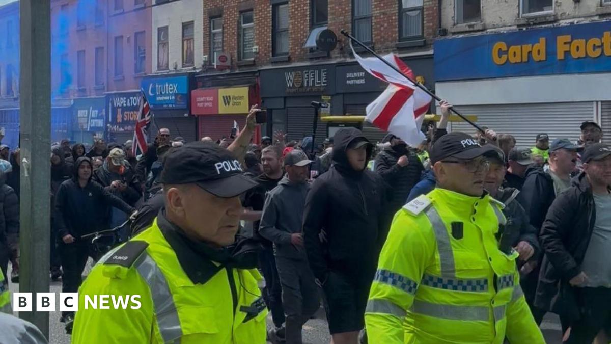 A large group of protesters, flanked by police officers in reflective yellow jackets, march along a street lined with shops, all of which have their shutters pulled down. Some protesters are carrying English and British flags, and some blue smoke from a flare is visible hanging in the air to the left.