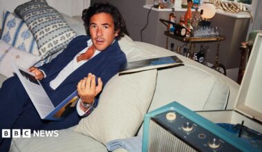 Jack Savoretti sitting on a white couch with white and colourful cushions. He is holding a vinyl record and is looking with surprise at the camera. There is a cart with alcohol behind him.