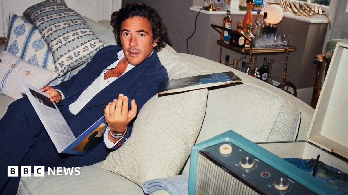 Jack Savoretti sitting on a white couch with white and colourful cushions. He is holding a vinyl record and is looking with surprise at the camera. There is a cart with alcohol behind him.