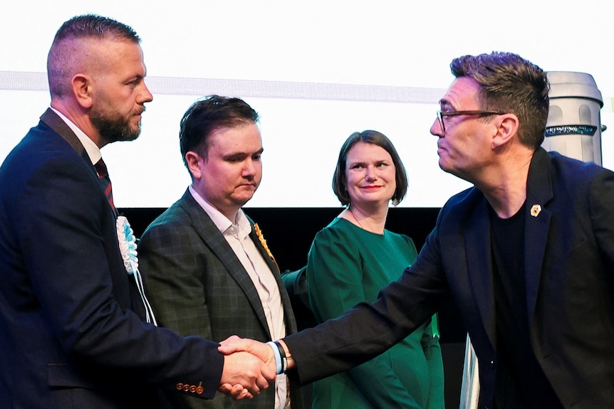 Two men shake hands, while a man and a woman look on in the background.