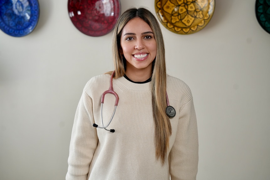 Ieesha Nasser in a cream knit, stethoscope around her shoulders and long hair, smiling.