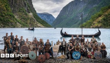 Norway players in Viking dress and with weapons and shields pose in front of a fjord with Viking boats
