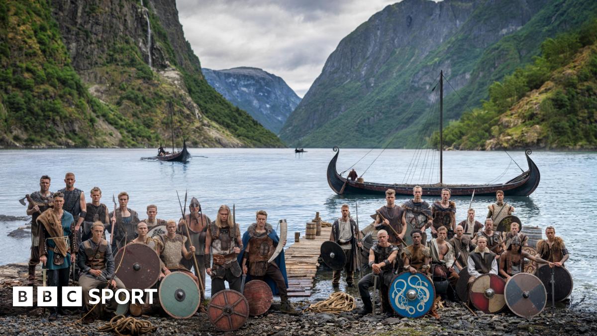 Norway players in Viking dress and with weapons and shields pose in front of a fjord with Viking boats