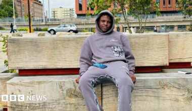 Panashe is sat on big wooden bench in Nottingham. He is wearing a lilac jumper and is looking towards the camera.