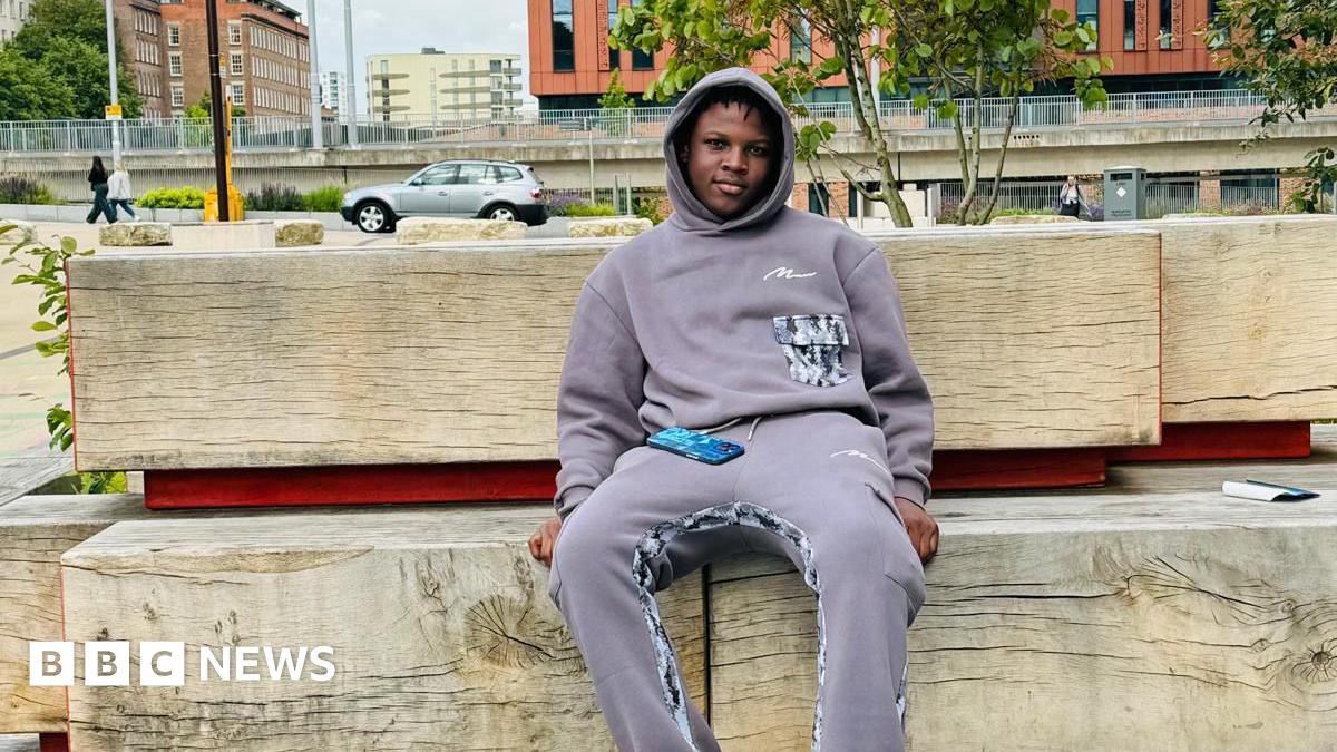 Panashe is sat on big wooden bench in Nottingham. He is wearing a lilac jumper and is looking towards the camera.