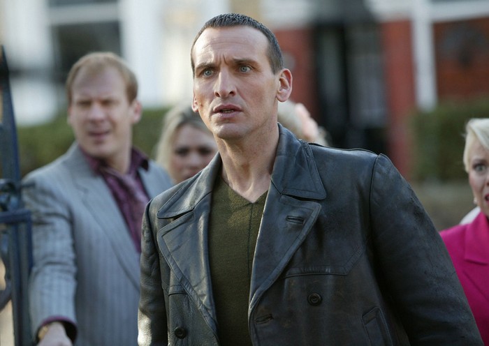 christopher-eccleston Christopher Eccleston as the Ninth Doctor in Doctor Who, wearing his iconic black leather jacket and green jumper, stands outdoors looking tense, with several people in the background also appearing alarmed.