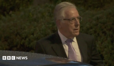 O'Brien has grey hair combed loosely back and wears glasses. He is wearing a dark suit jacket and a lilac tie.