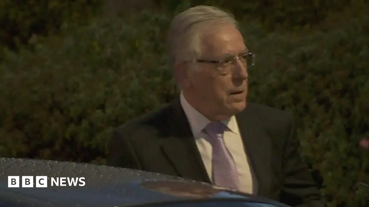 O'Brien has grey hair combed loosely back and wears glasses. He is wearing a dark suit jacket and a lilac tie.