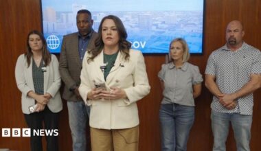 A woman in a white blazer stands in the foreground with a cell phone in her hands as she speaks. In the background of her are four other individuals leaning up against a wooden wall.