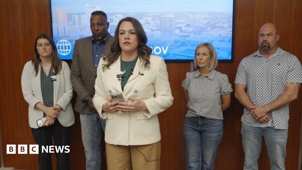 A woman in a white blazer stands in the foreground with a cell phone in her hands as she speaks. In the background of her are four other individuals leaning up against a wooden wall.