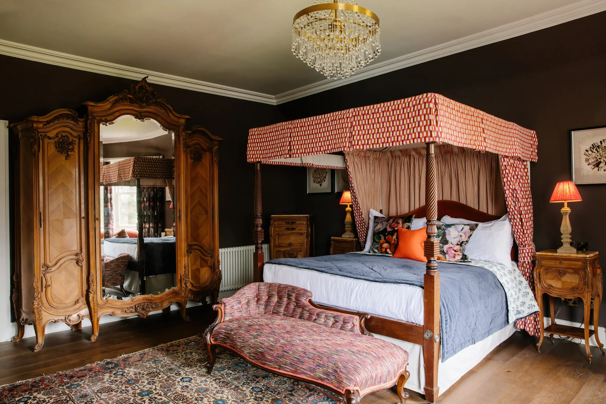 A bedroom with a dark brown accent wall, a four-poster bed with a red and white canopy, a red patterned fainting couch, and a large wooden wardrobe with a mirror.