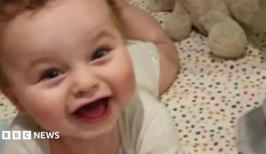 A smiling baby lies on a cot with his head up and rosy cheeks