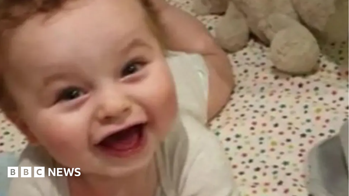 A smiling baby lies on a cot with his head up and rosy cheeks